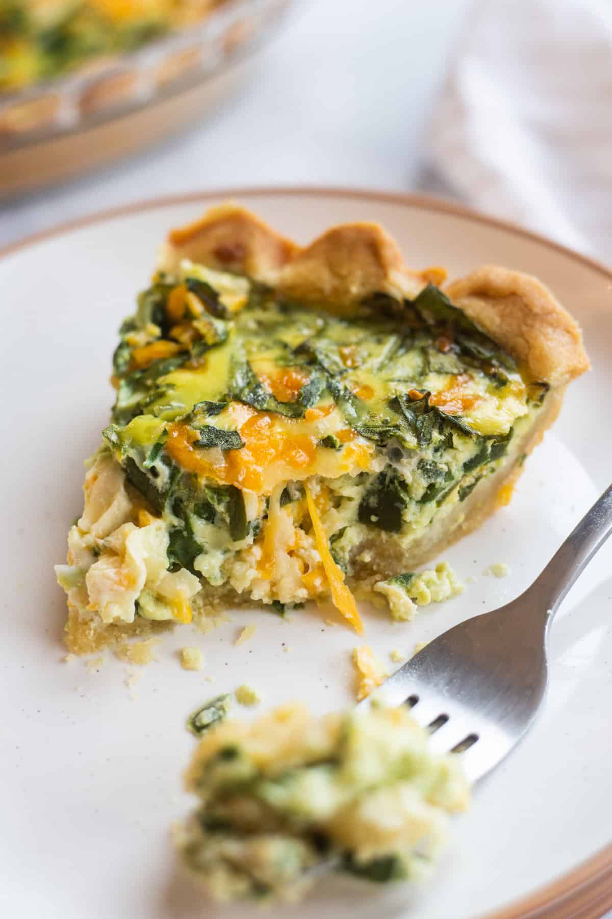 slice of spinach cheese quiche with a bite taken out of it served on a white round plate