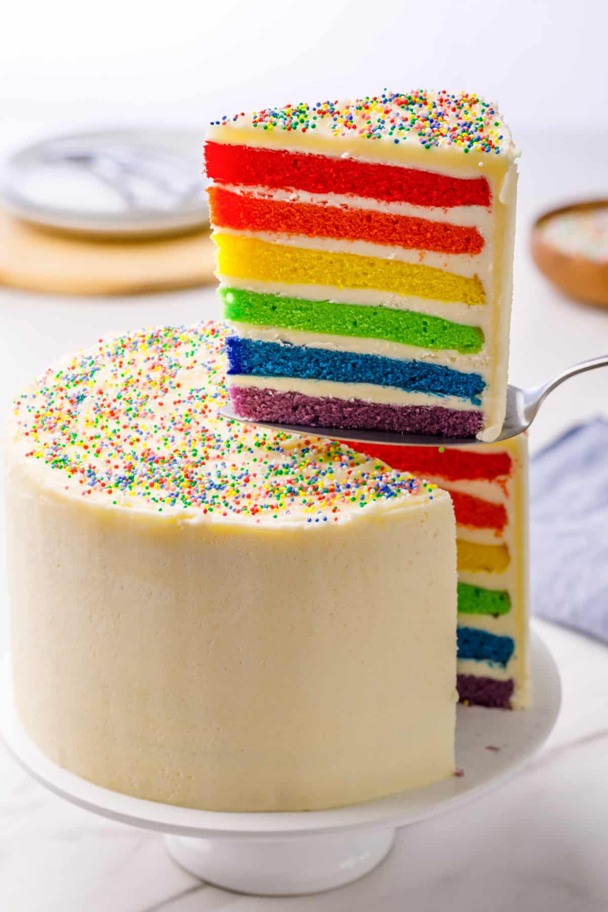 rainbow cake served on a cake stand with a slice of cake being taken out with a cake spatula