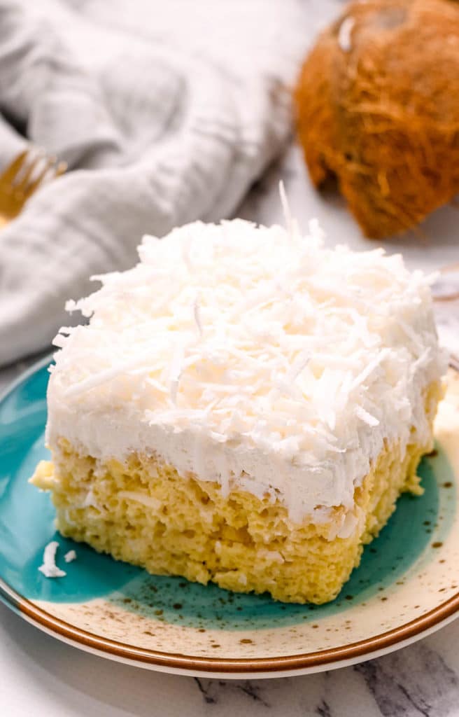 A slice of coconut cream cake on a plate with coconut flakes.