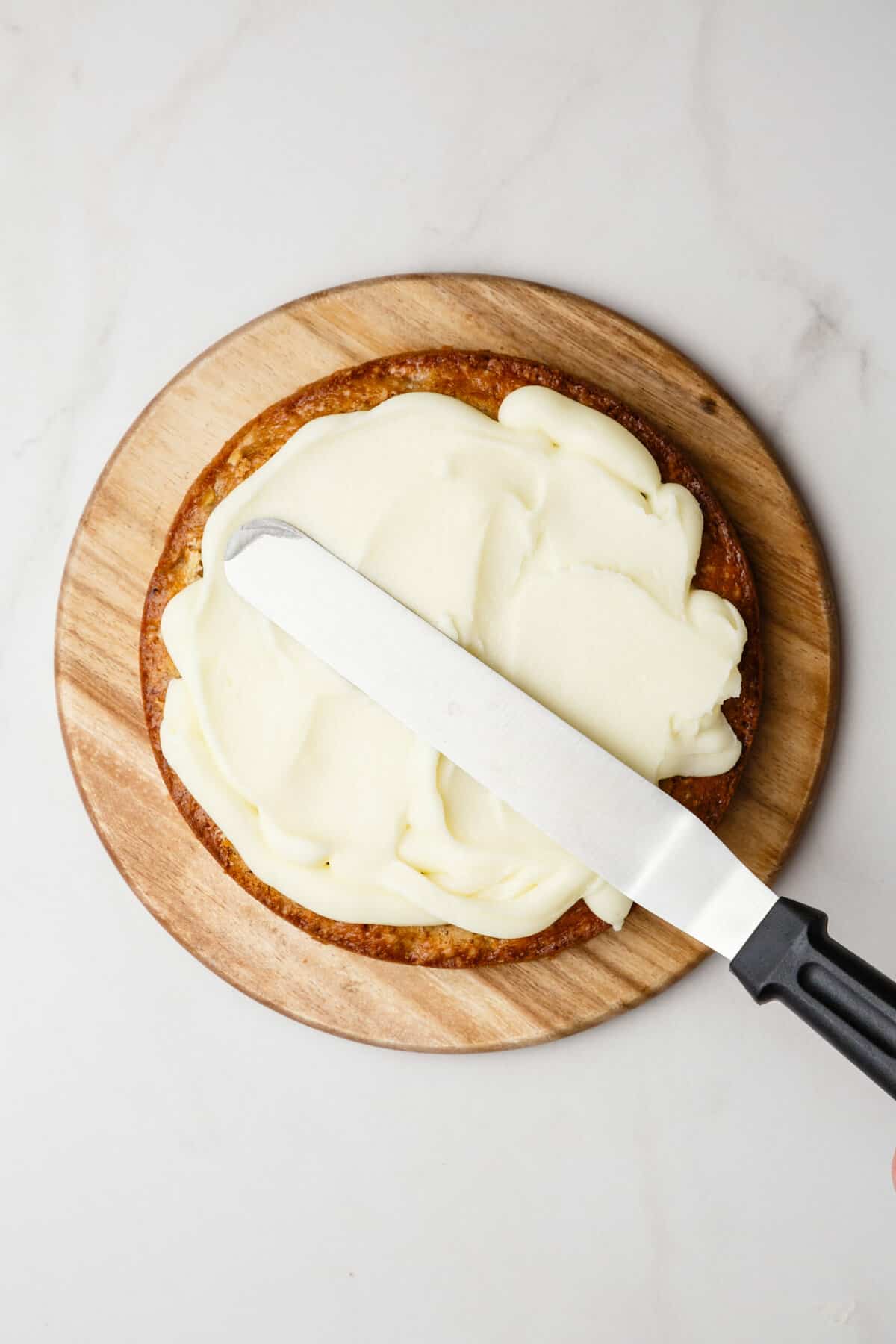 top down image of an off set spatula frosting a vanilla cake sitting on a round wooden board. 