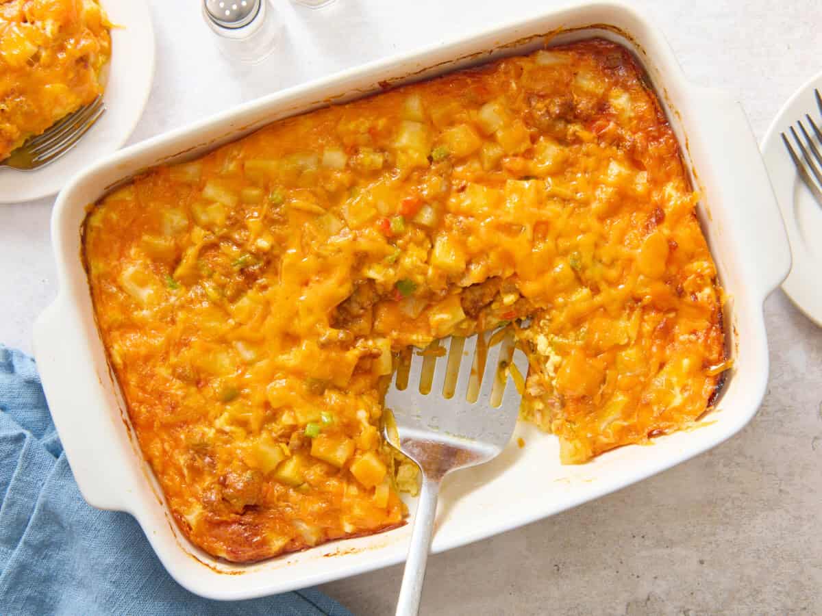 Spatula in casserole dish of overnight breakfast bake with egg and sausage.