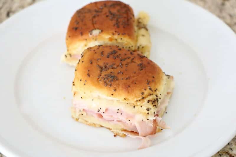 White plate with two ham and cheese sliders on mini rolls with poppy seeds on top.