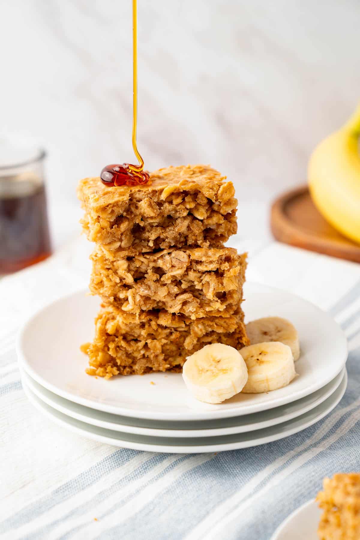 A stack of three peanut butter banana oatmeal bars sitting on a white place on a white counter. Honey is drizzling over the top of the bars.