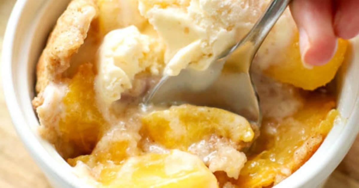 A spoon digging into a bowl of peach cobbler topped with ice cream. 