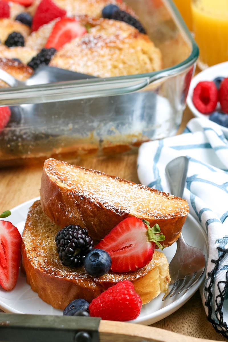 Overnight baked french toast casserole slices with berries stacked on a plate and the french toast casserole in the pan in the background
