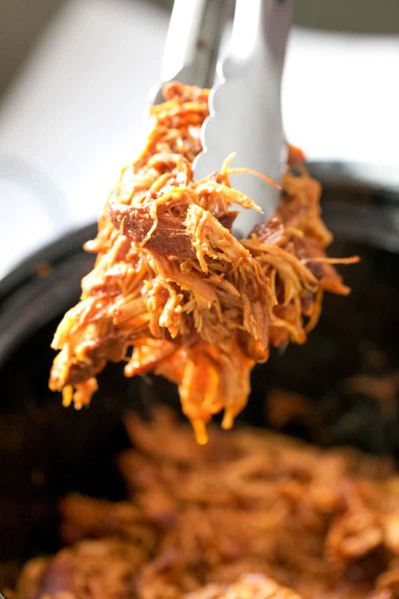 Tongs grabbing a pinch of bbq chicken out of the slow cooker.