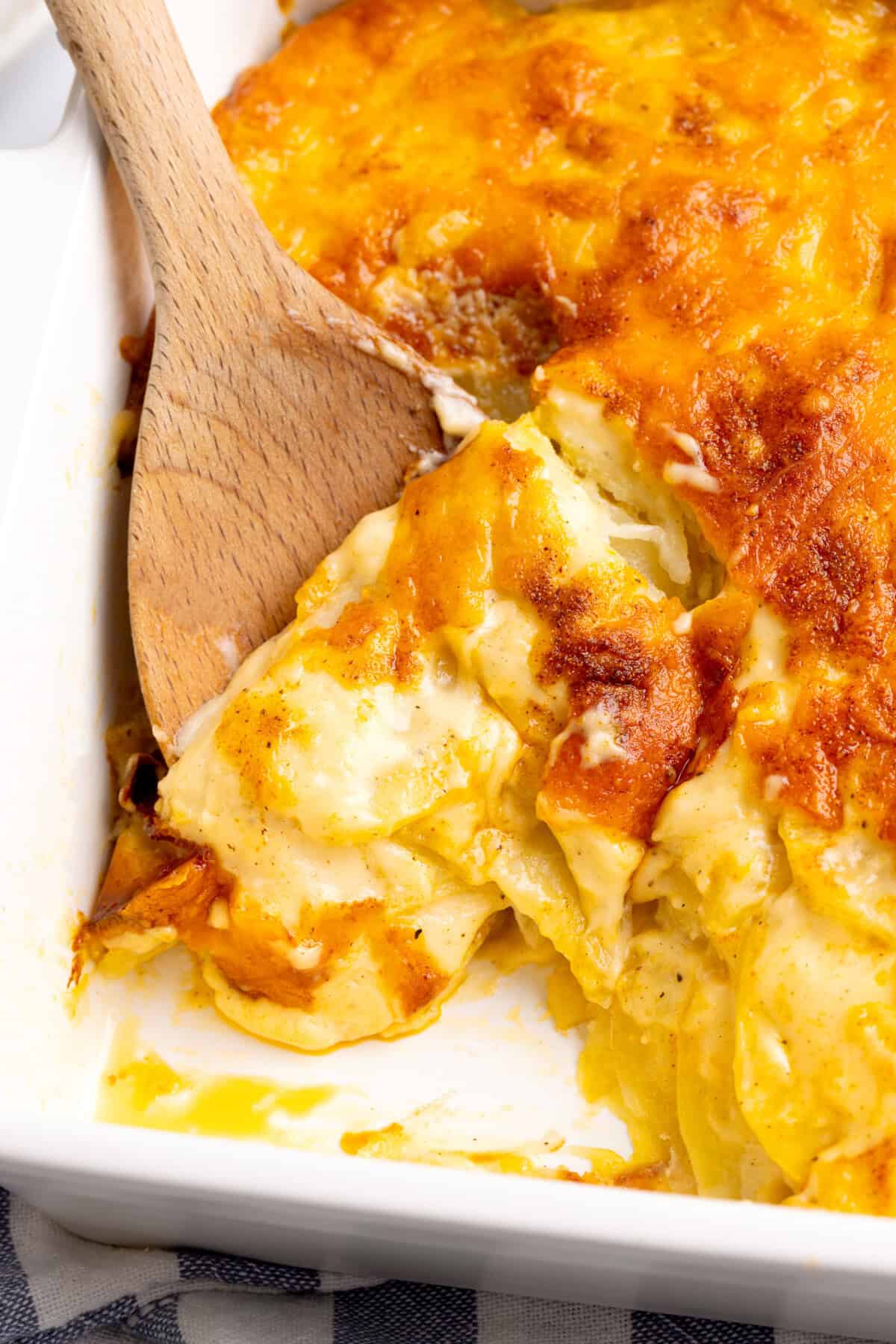 Wooden spoon scooping cheesy scalloped potatoes from pan.