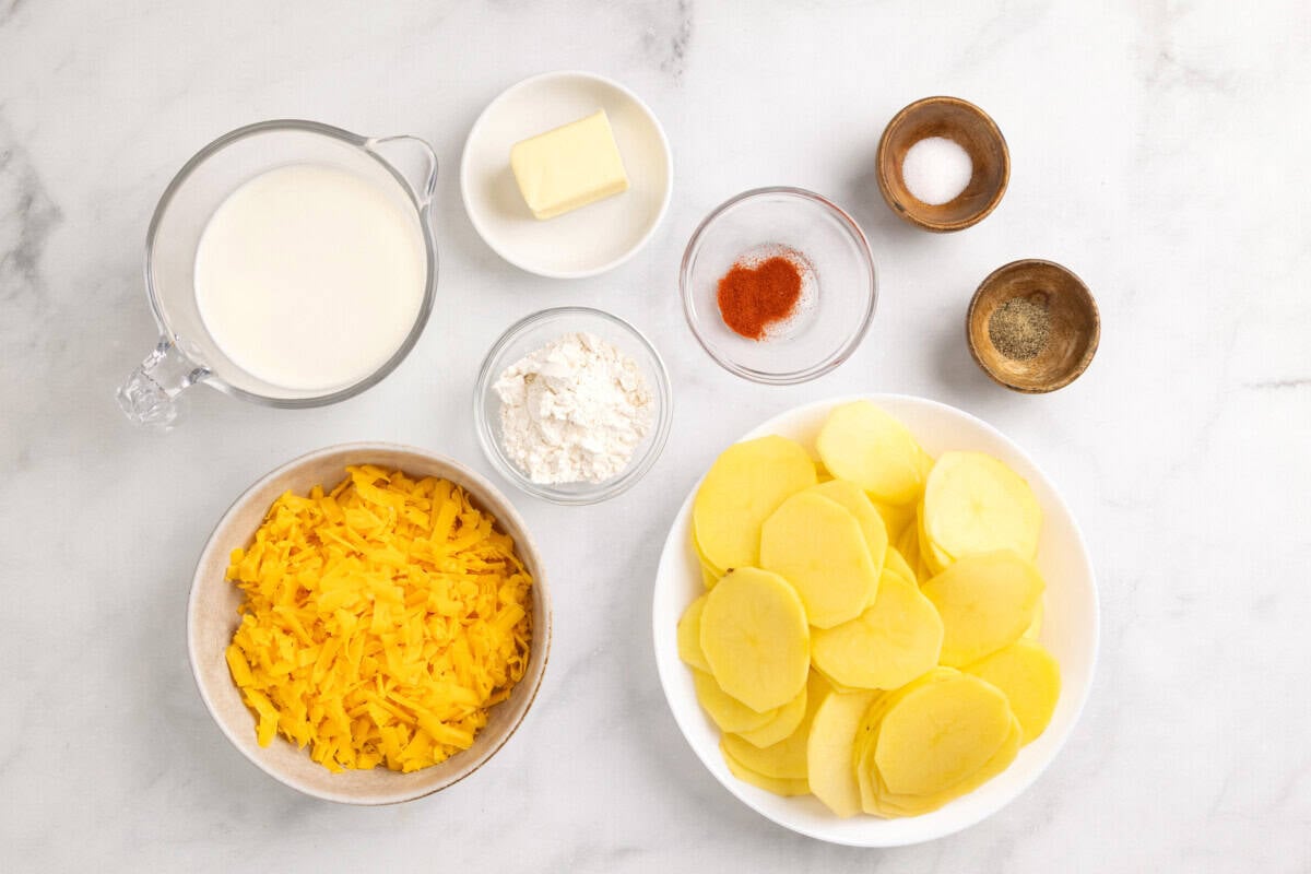 Ingredients for cheesy scalloped potatoes, including potatoes, cheese, milk, flour, paprika, butter, salt, and pepper.
