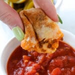 Hand dipping piece of taco stick into marinara sauce held in small white bowl