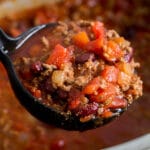 A black ladle full of homemade crock-pot chili with ground beef and beans hovers over a grey slow-cooker full of chili.