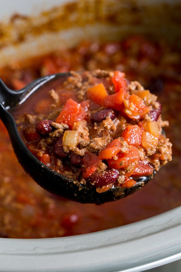 A black ladle full of homemade crock-pot chili with ground beef and beans hovers over a grey slow-cooker full of chili. 