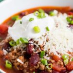 Close up image of crock pot chili in a white bowl with a silver spoon. This ground beef and bean chili is topped with shredded cheddar cheese, sour cream, and fresh scallions.