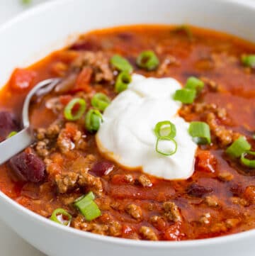 Image of easy slow cooker ground beef chili with beans in a white bowl, topped with sour cream and fresh scallions. A silver spoon is dipped in the bowl.