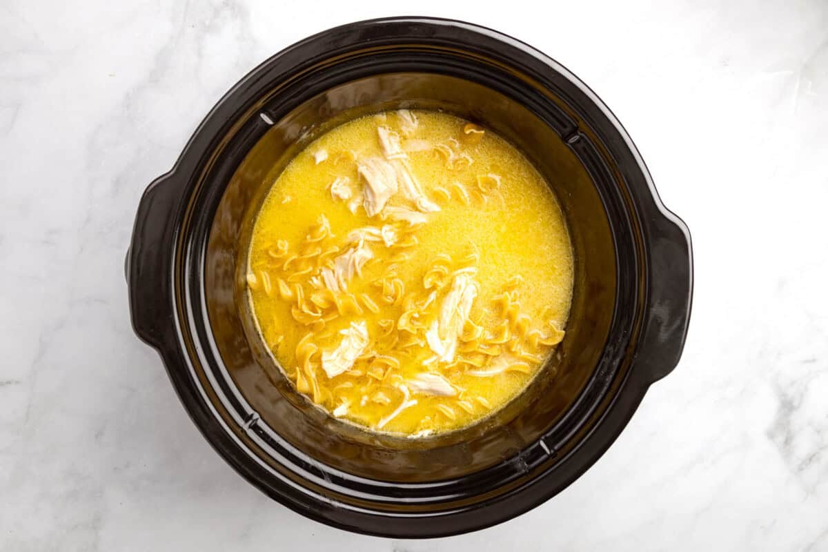 The shredded cooked chicken in the broth with the noodles in the crockpot.