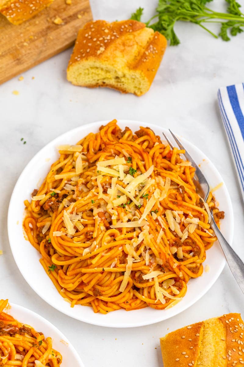 Spaghetti in red meat sauce on a white plate, with bread in the background.