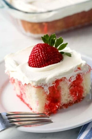 A piece of strawberry jello poke cake sits on a white plate with a fork on the side.