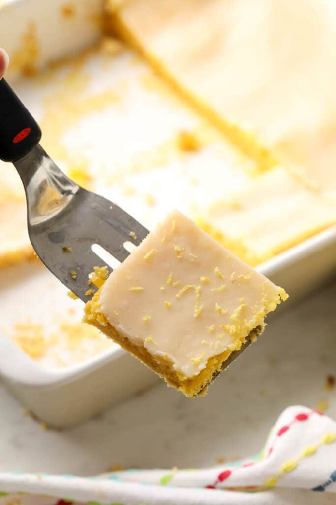 a cut lemon bar topped with lemon zest being held on a spatula. 