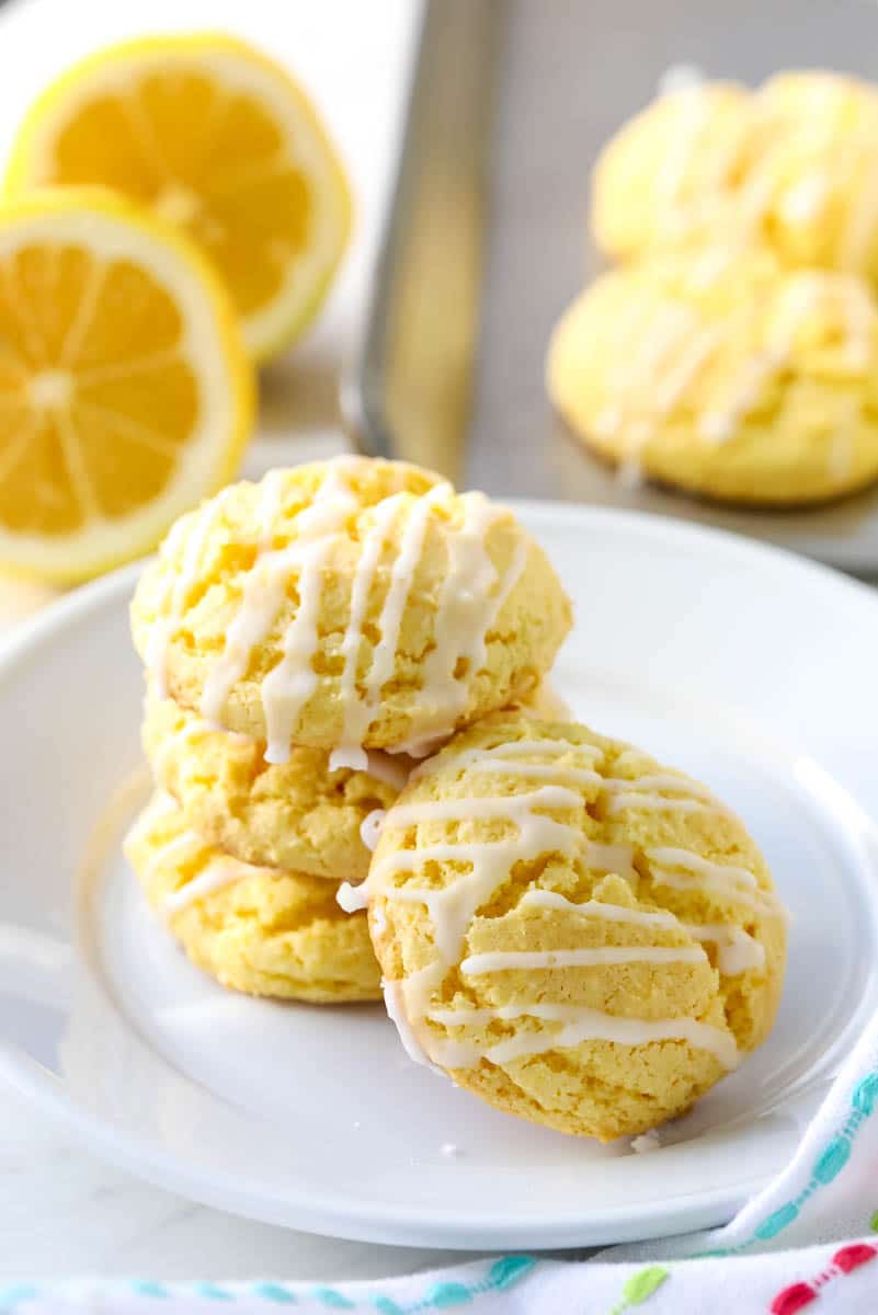 Lemon cookies drizzled with a glaze topping stacked on a white plate with cookies on a pan and lemons in the background.