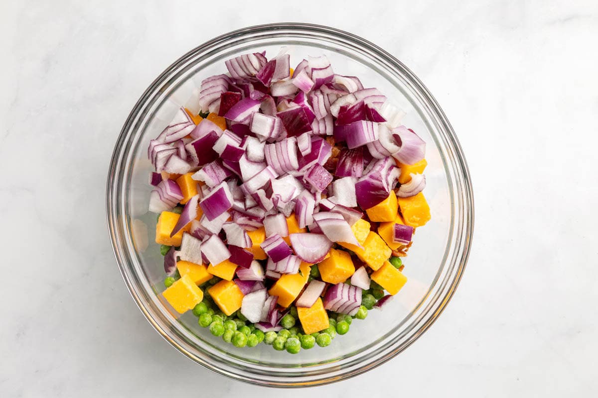 The peas, bacon, red onion, and cheddar cheese in a bowl before mixing together.