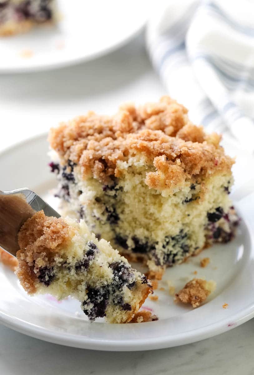 Blueberry buckle coffee cake slice closeup on a plate showing inside of cake with blueberries and struesel topping, with fork scooping a bite.