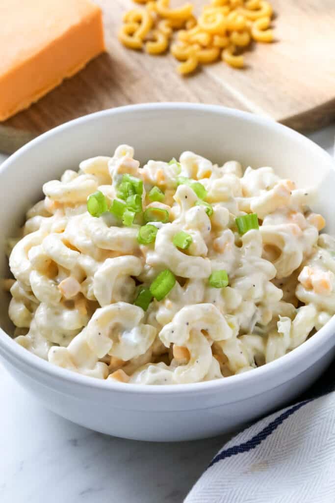 Macaroni salad in a white bowl set on a marble counter alongside a white dishrag, a block of cheddar cheese, and elbow macaroni.