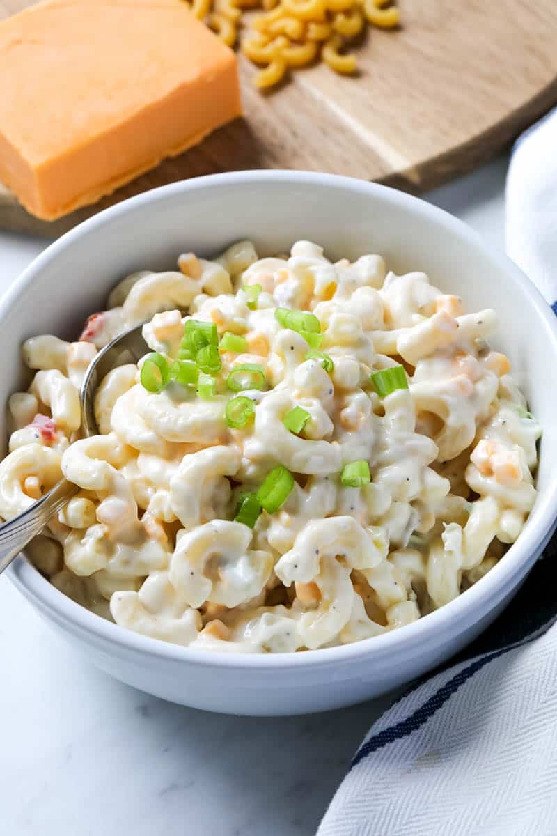 Macaroni salad in a white bowl with a metal spoon, set on a marble counter alongside a white dishrag, a block of cheddar cheese, and elbow macaroni.