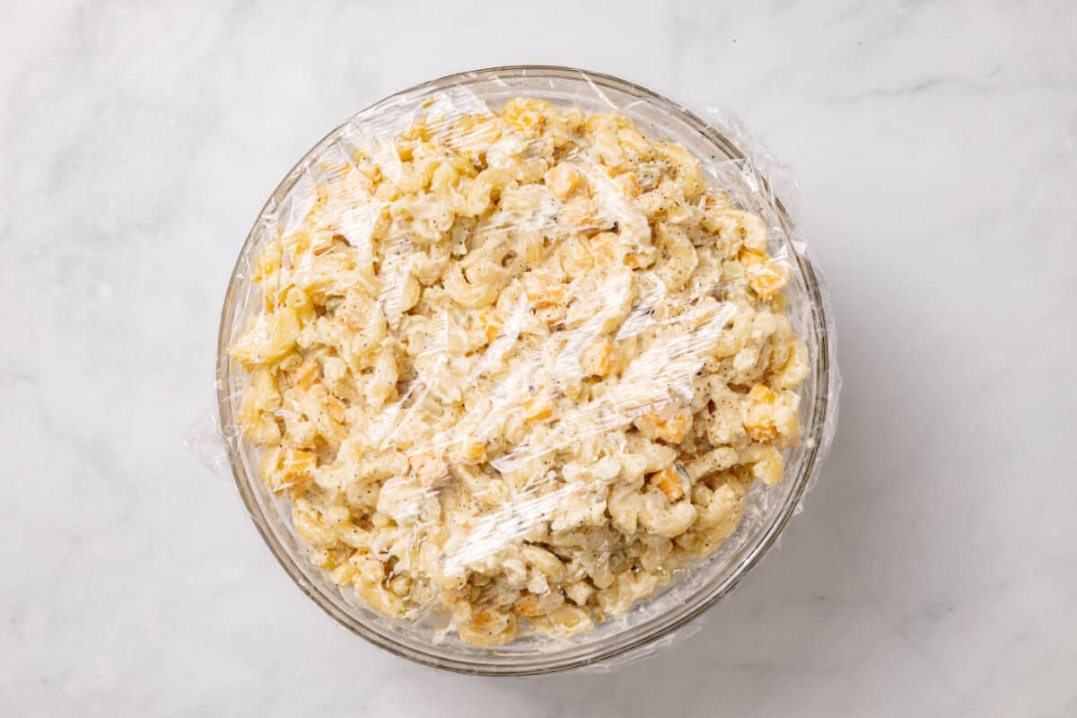 Macaroni pasta salad in glass bowl with plastic wrap cover.