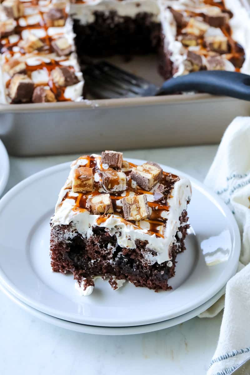 snickers poke cake on a plate by a 13x9 pan of cake