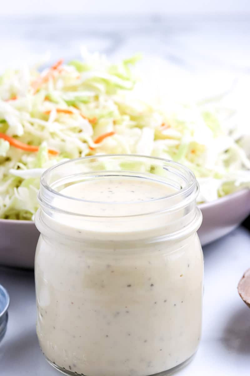 jar of coleslaw dressing in front of a white bowl filled with coleslaw