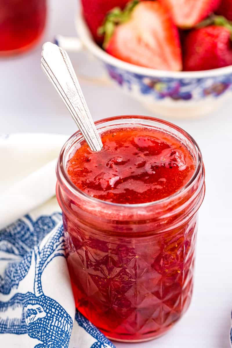 open jar of strawberry rhubarb jam with a spoon in it, in front of a bowl of sliced strawberries