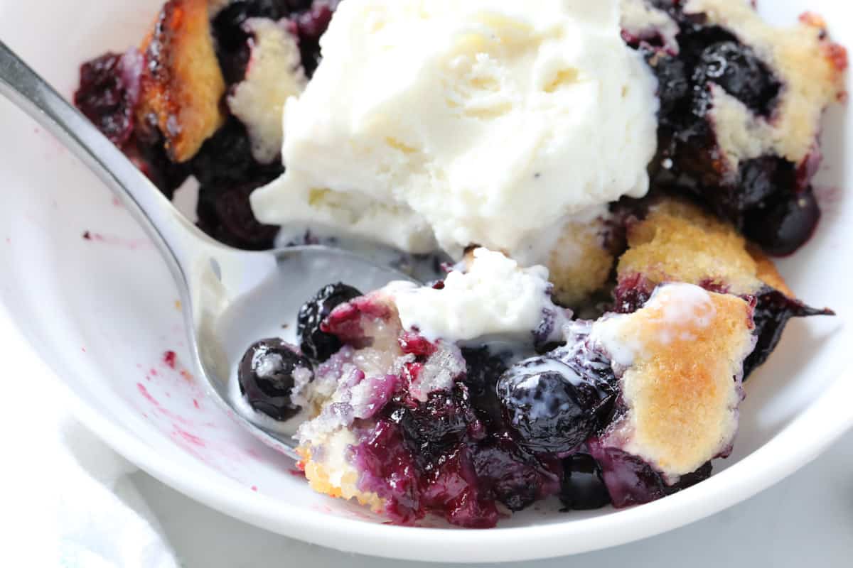 A spoon lifting a bite of blueberry cobbler topped with ice cream from a bowl. 