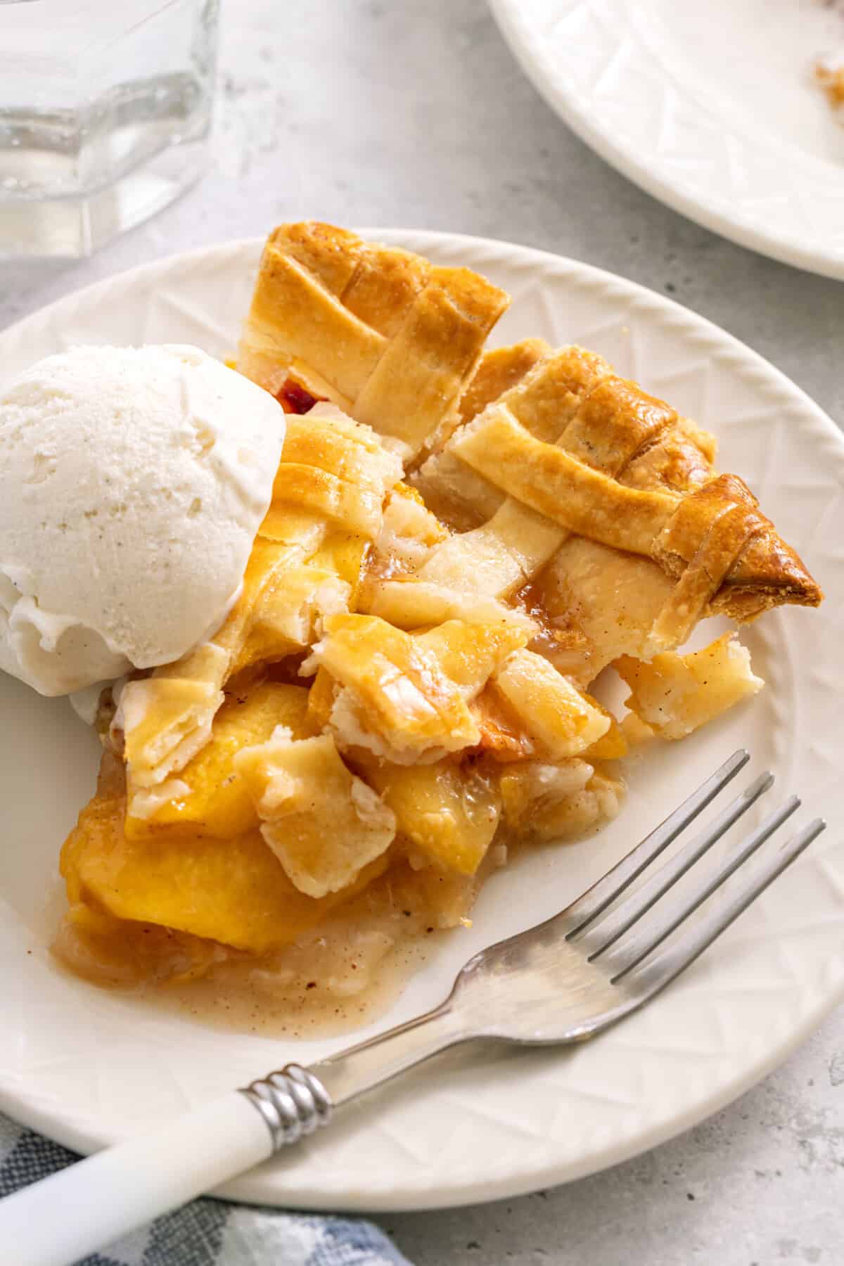 Slice of homemade peach pie on plate with fork and scoop of vanilla ice cream.