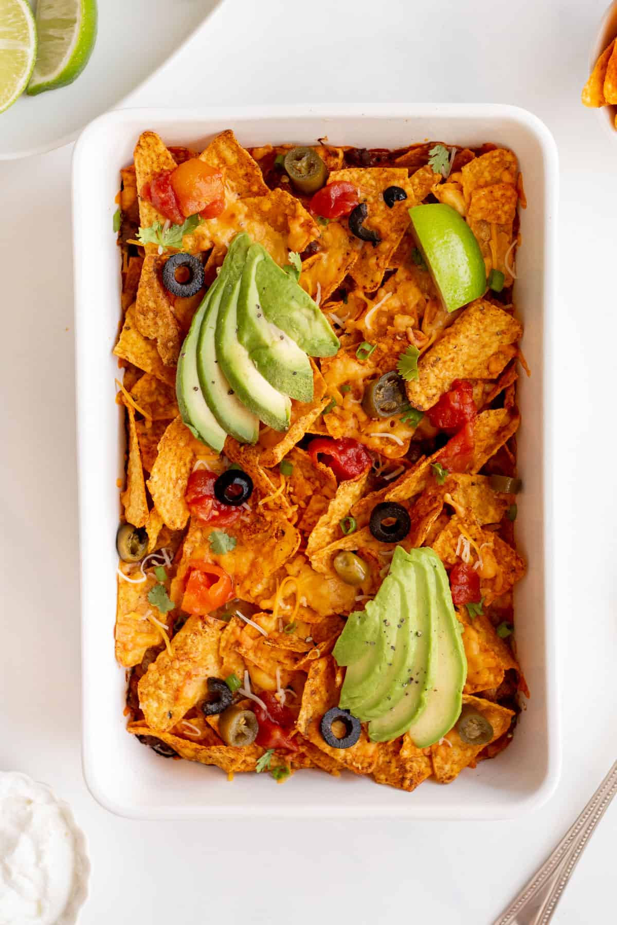 doritos casserole in a white rectangle dish, topped with sliced black olives, tomatoes, jalepenoes, avocados, cilantro, shredded cheese, and lime