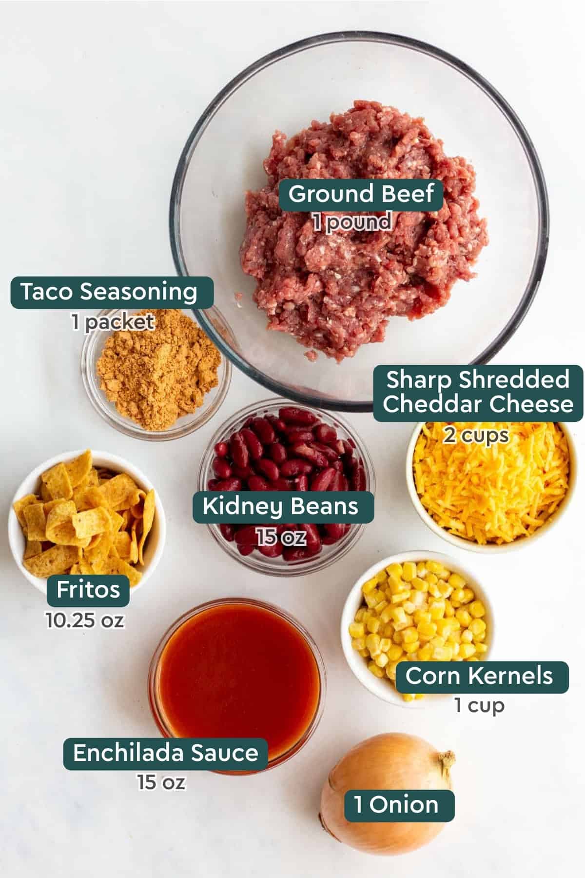 Ground beef, onion, cheese, beans, tomato sauce, taco seasoning, and Fritos laid out on kitchen counter.