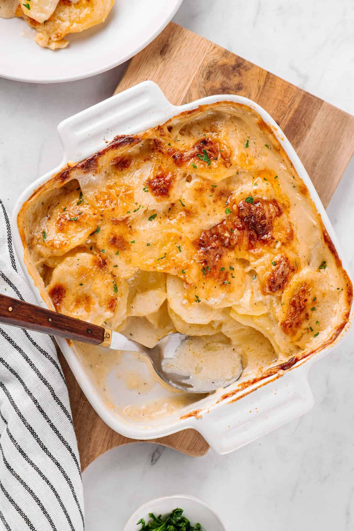 potatoes au gratin served in a white square casserole dish sitting on a wood cutting board with a spoon in the dish