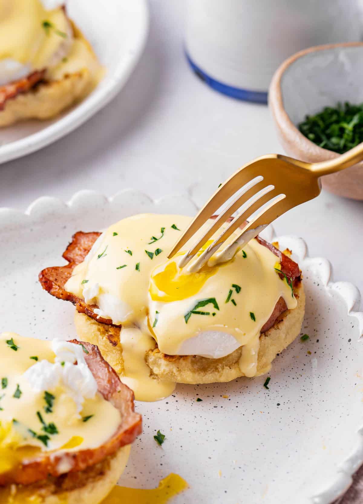 eggs benedict with ham, poached eggs, and homemade hollandaise sauce served on a speckled and scalloped plate with a gold fork cutting into the egg
