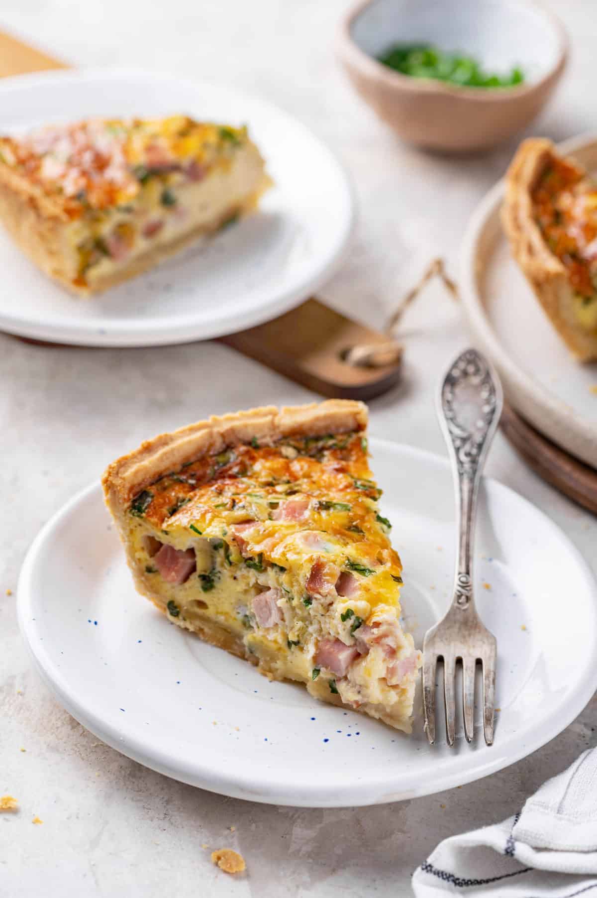 slice of ham and cheese quiche served on a white round plate with a silver fork