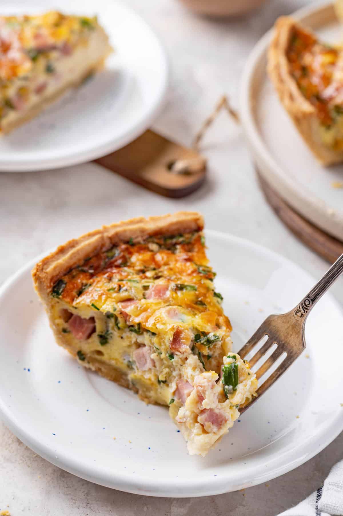 slice of ham and cheese quiche served on a white speckled plate with a forkful of the quiche