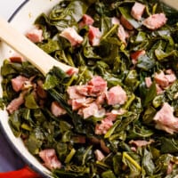 Collard greens with ham hocks and bacon being served on a wooden spoon.