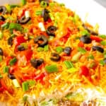 7 layer bean dip served in a casserole dish topped with guacamole tomatoes cheese green onions and black sliced olives