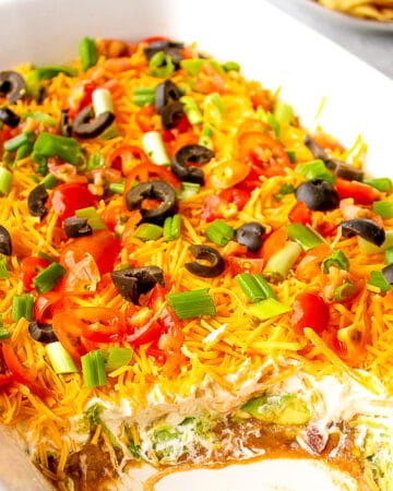 7 layer bean dip served in a casserole dish topped with guacamole tomatoes cheese green onions and black sliced olives