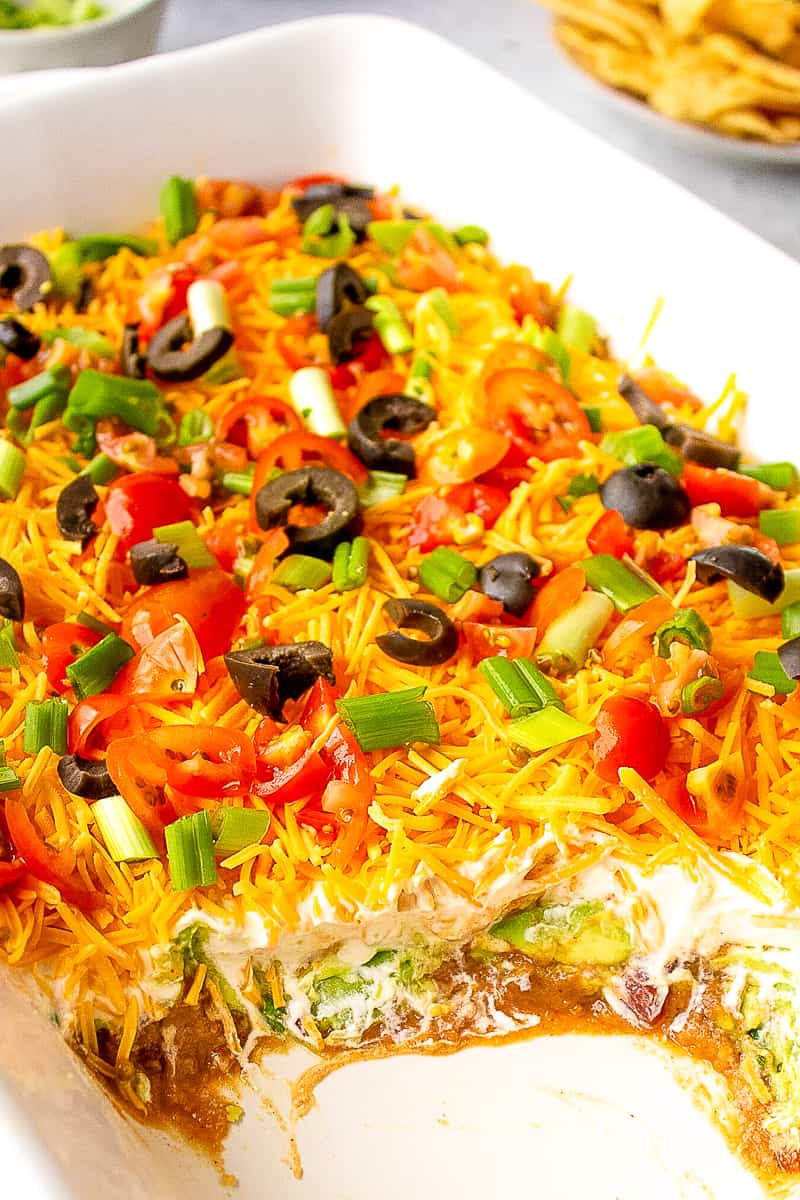 7 layer bean dip served in a casserole dish topped with sour cream, guacamole, shredded cheddar cheese, green onions, and black olives