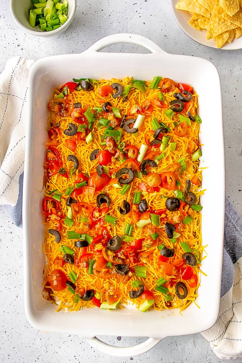7 layer bean dip served in a casserole dish topped with sour cream, guacamole, shredded cheddar cheese, green onions, and black olives