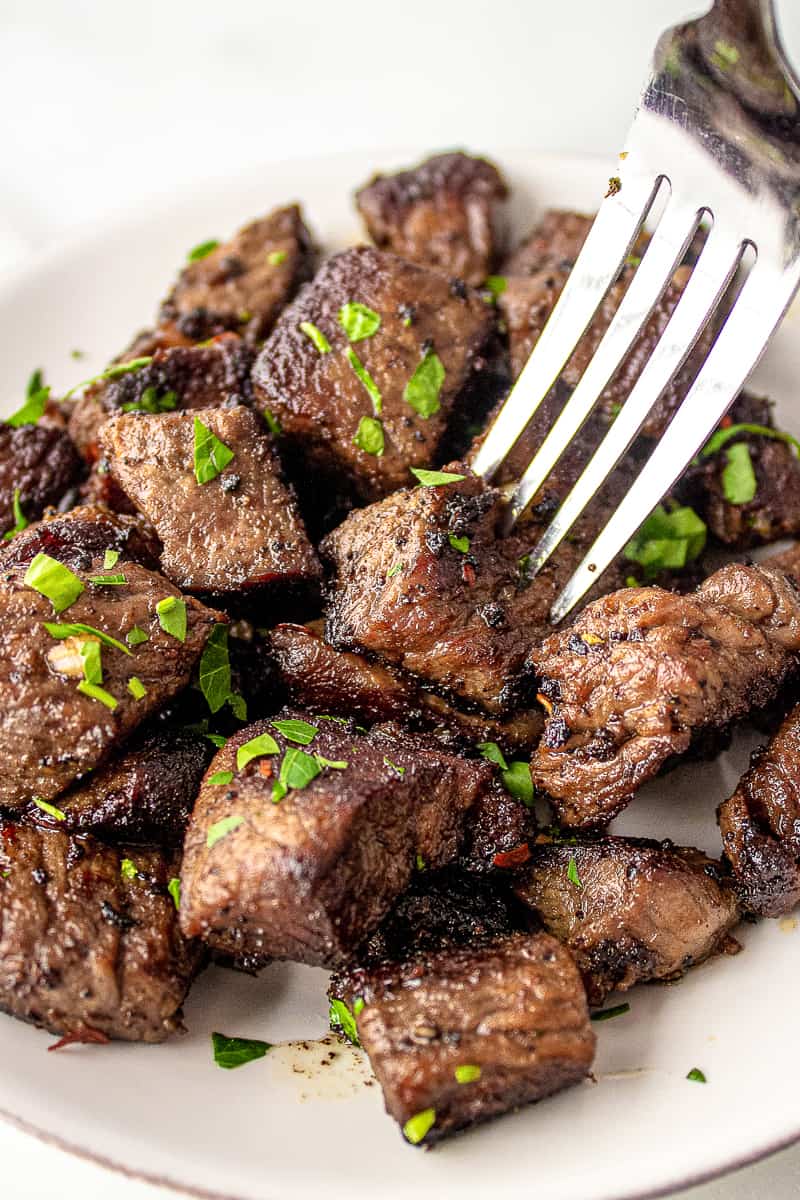 garlic butter steak bites garnished with fresh chopped parsley served on a white plate with a silver fork digging into the a steak bite