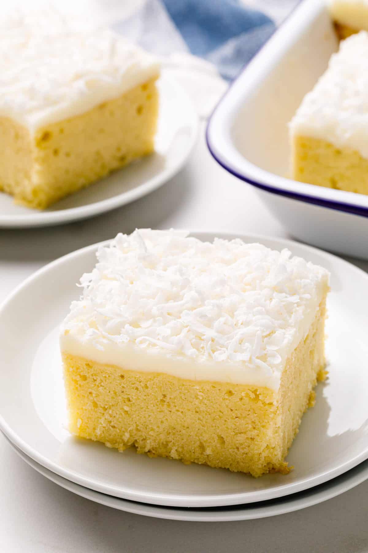 square slice serving of coconut cake with coconut cream frosting on a white round plate