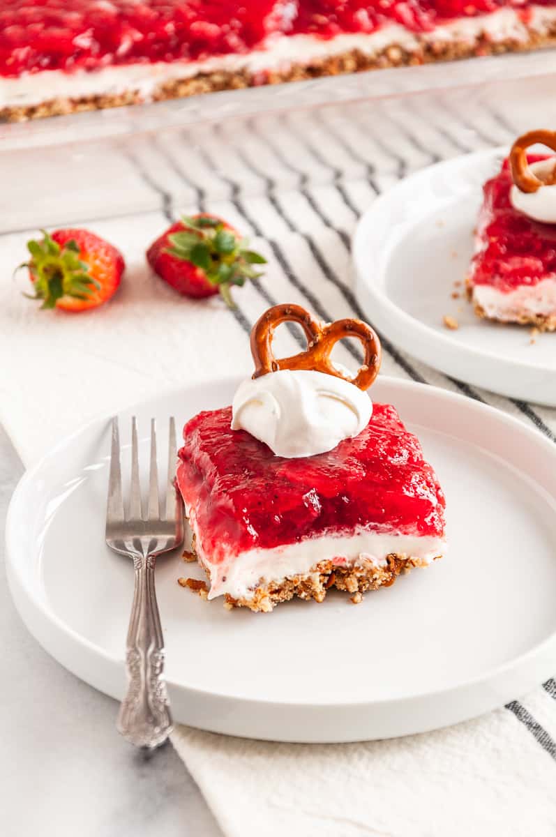 square serving of strawberry pretzel dessert topped with a dollop of whipped cream and a pretzel served on a white plate with a silver fork