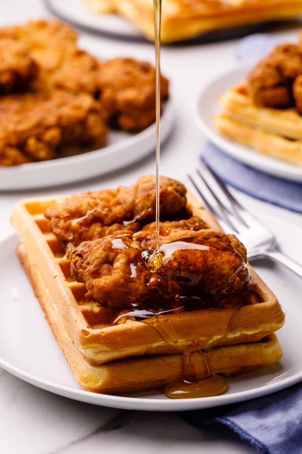 chicken and waffles with syrup being poured on and served on a white round plate with a silver fork