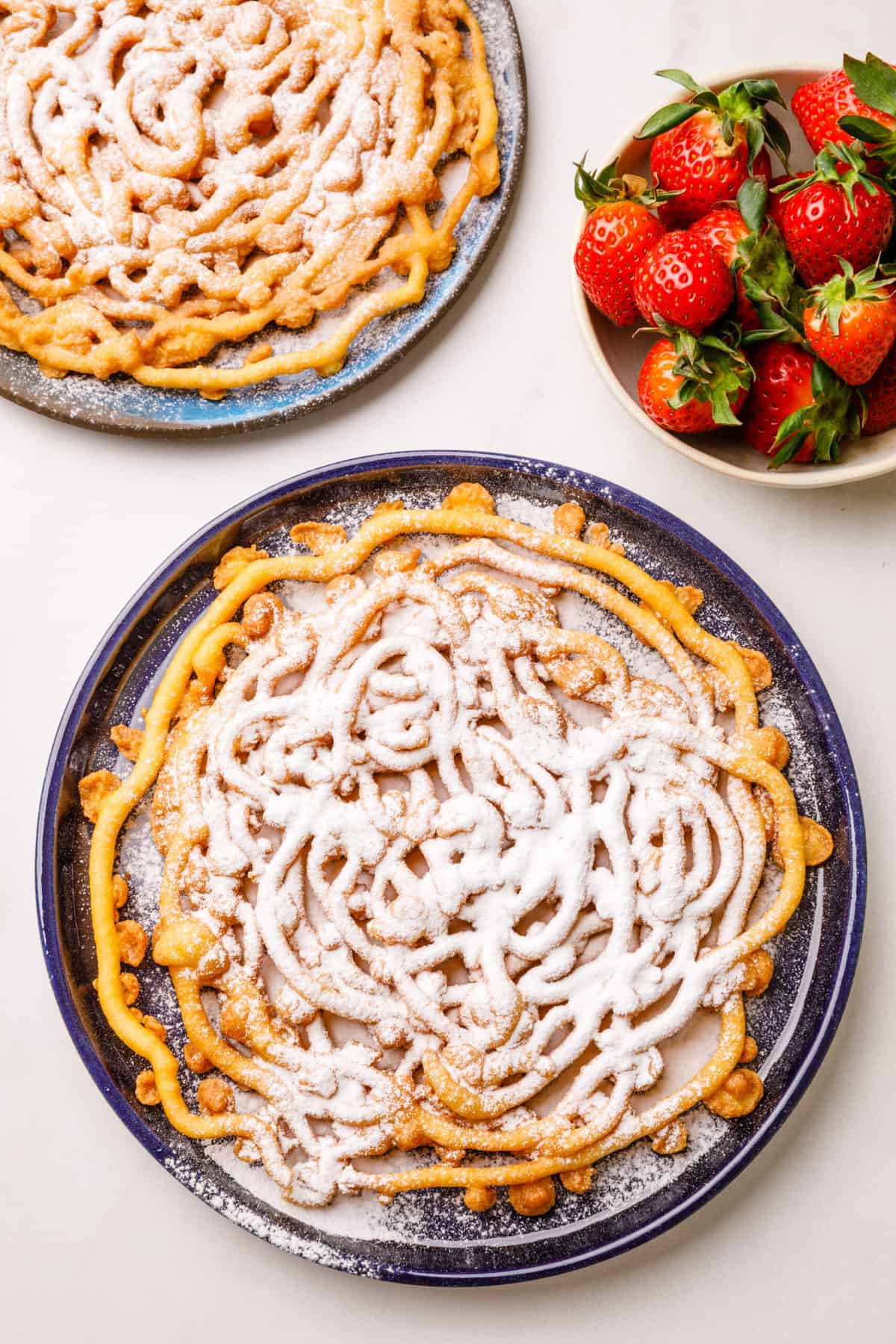 top down view on funnel cake served on a blue round plate with a bowl of strawberries to the side