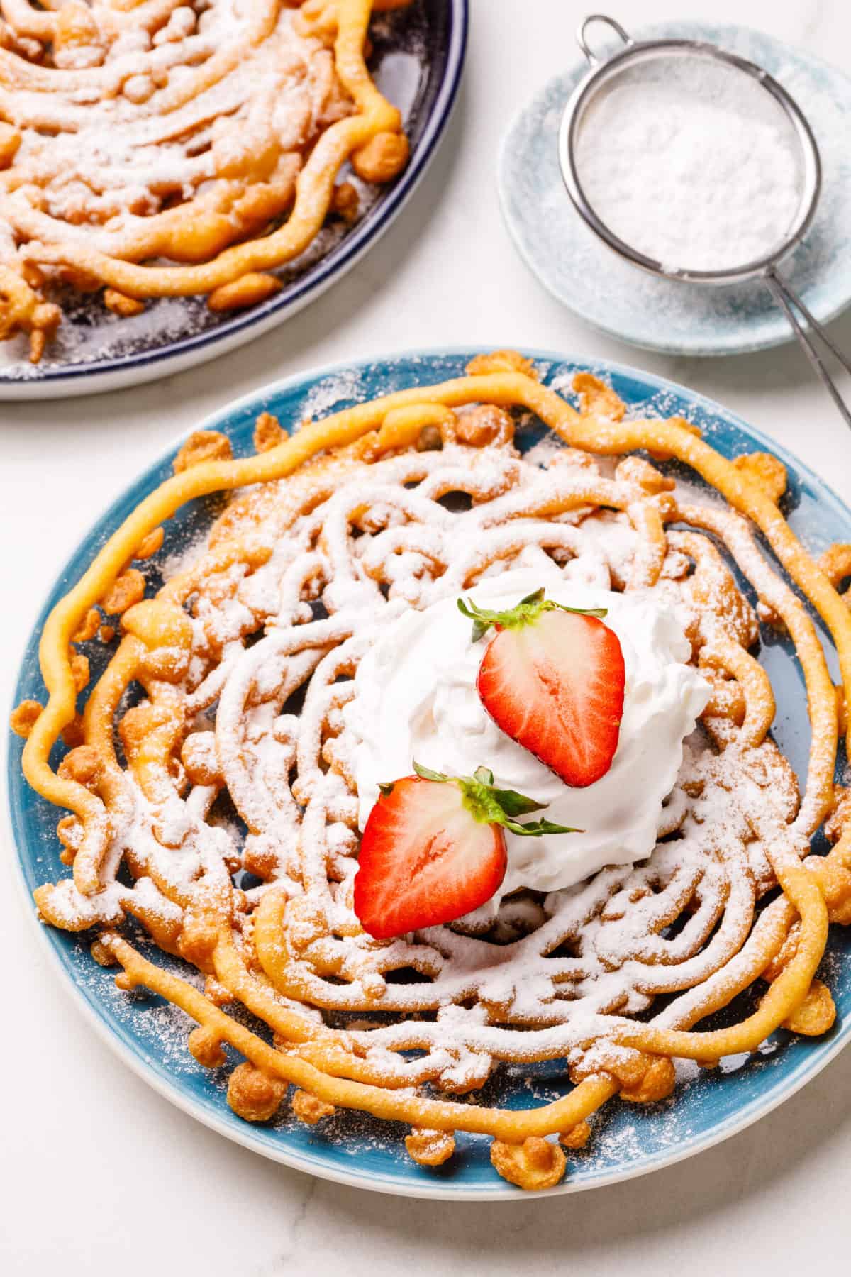 homemade pancake mix funnel cake topped with powdered sugar, whipped cream, and fresh halved strawberries served on a blue round plate