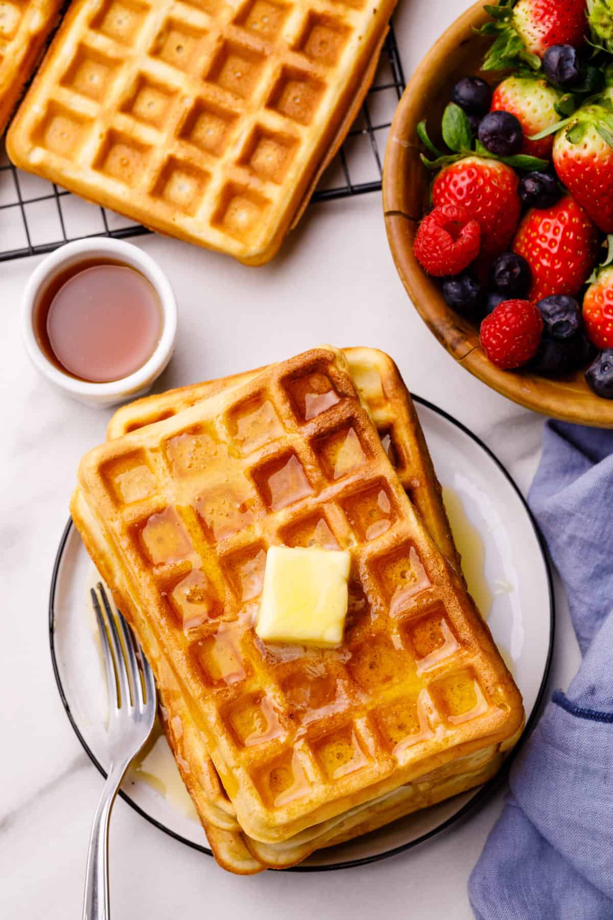 waffles stacked on a plate topped with a slab of butter and syrup with a silver fork
